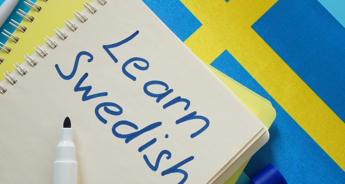 Bild: Öppen anteckningsbok med texten "Learn Swedish" och en svensk flagga i bakgrunden.
