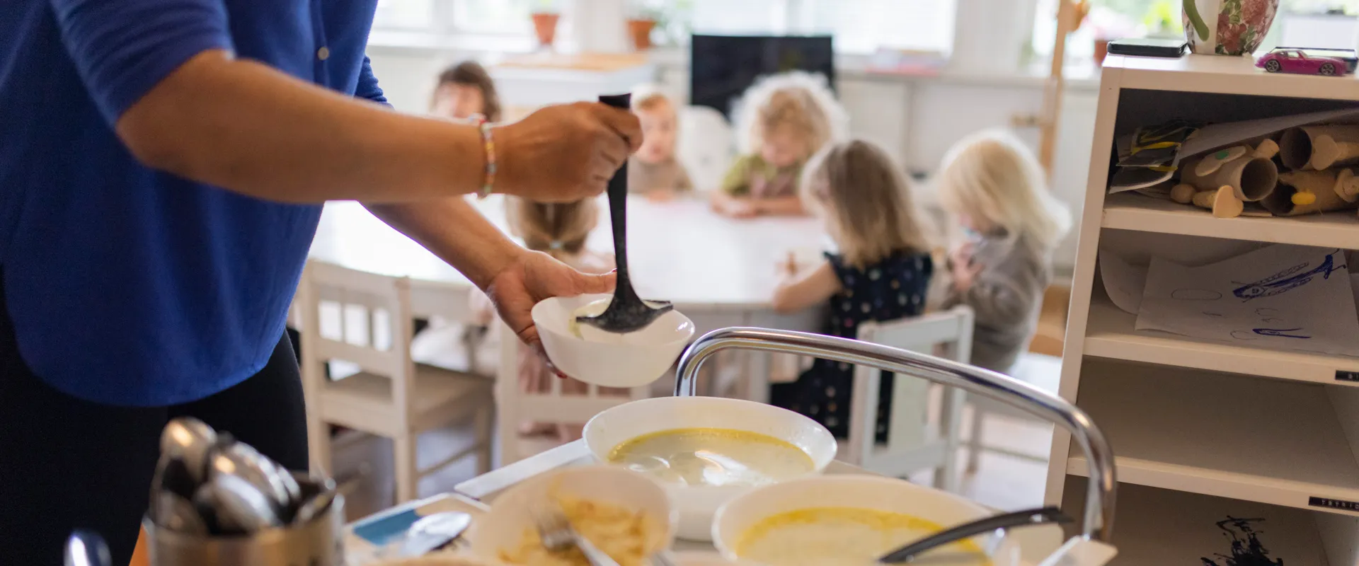 En kvinna som häller mat från en skål till en annan skål. Barn i förskoleålder sitter runt ett matbord i bakgrunden.